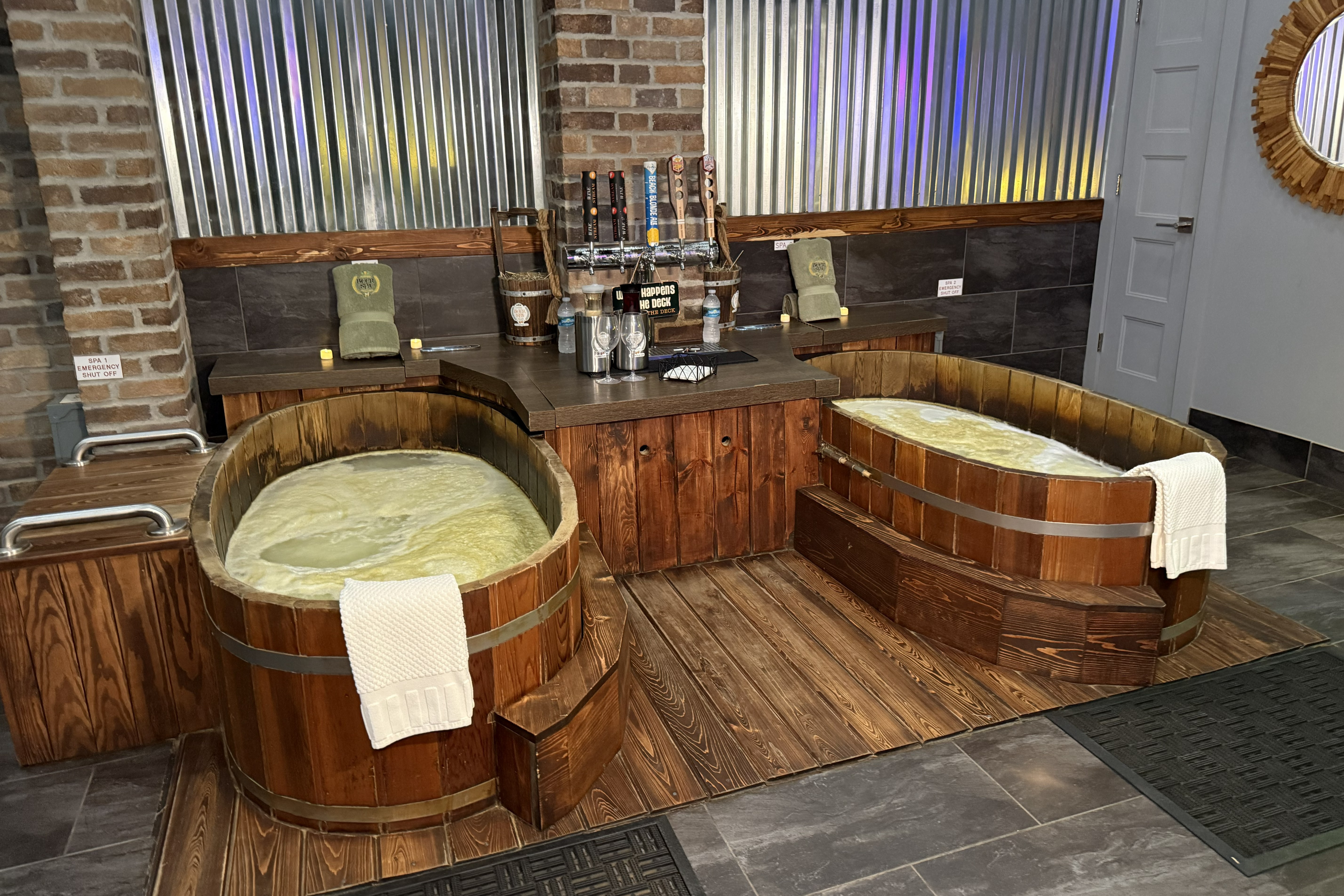 Two large wooden soaking tubs with yellow foamy water sitting in front of a bar and a wall decorated in aluminum. 