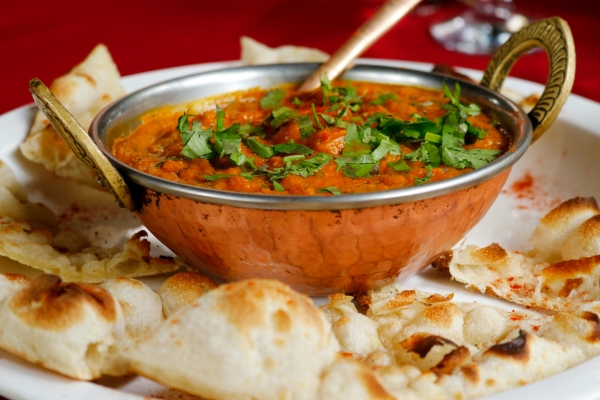 A small metal bowl of a sauce-based Indian dish sprinkled with fresh herbs and surrounded by sliced naan.