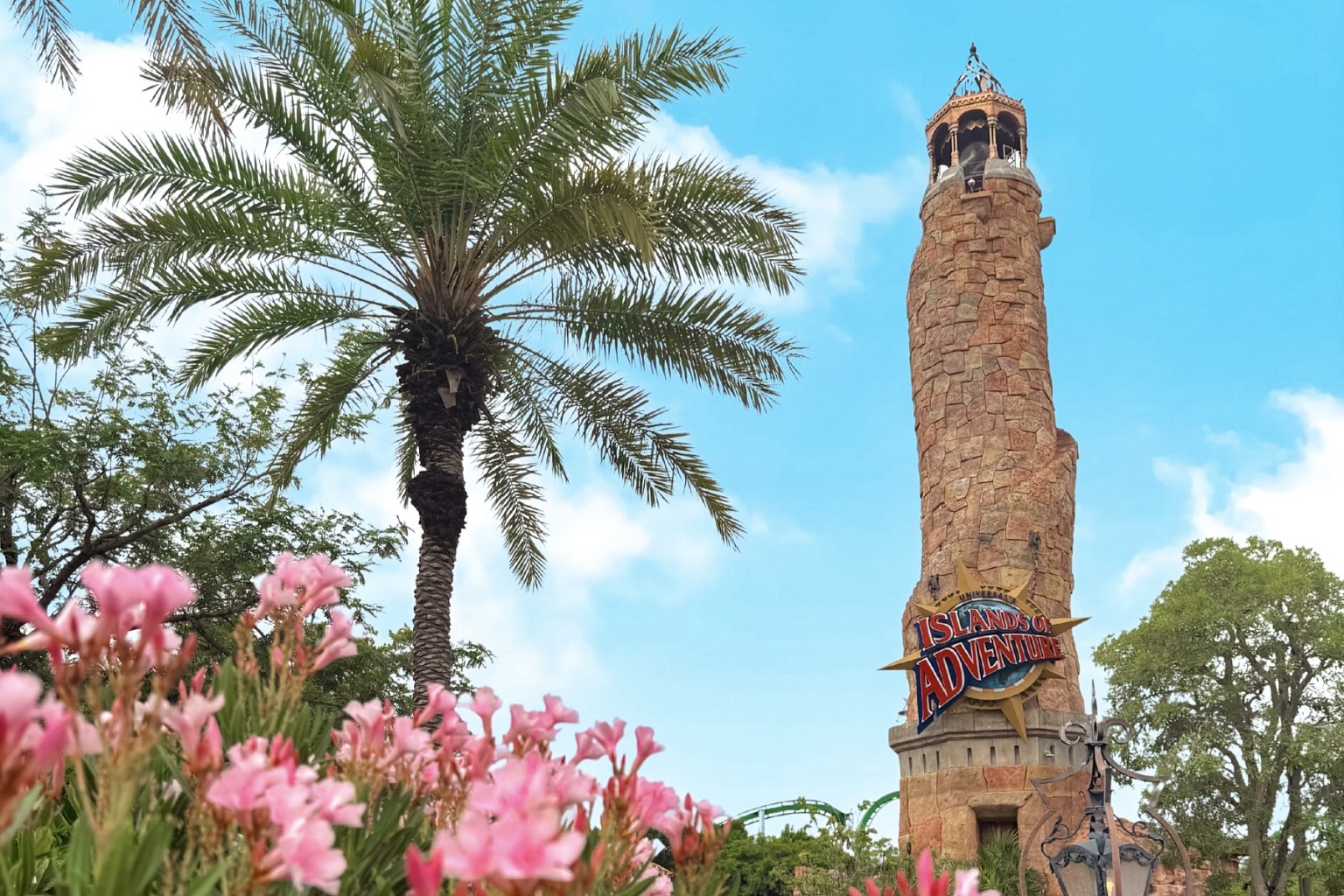 A stone tower with a sign that reads Islands of Adventure next to a palm tree with pink flowers in the foreground. 