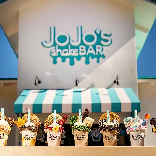A lineup of opulent milkshakes staged outside the front of a building with a sign reading JoJo's ShakeBAR and a blue and white striped awning.