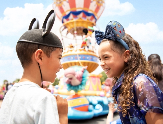 A boy and a girl in Mickey Ears grin at each other while a large colorful parade float stands out of focus in the background. Magic Kingdom is a great choice if you only have one day to visit Orlando theme parks.