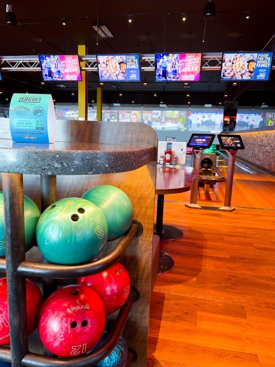 A rack with colorful bowling balls against a background of bowling lanes with colorful tv screens.