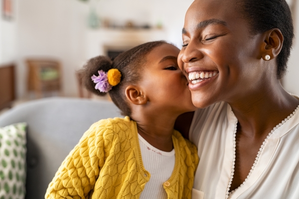 A girl in a yellow cardigan with pigtails kisses a woman in a white shirt on the cheek.