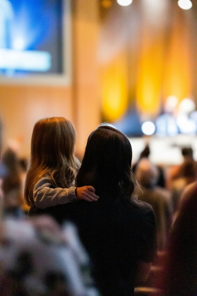 The back of a woman holding a young girl as they face a lit stage togehter.