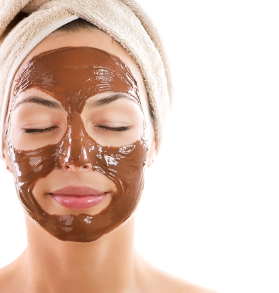 A woman with her eyes closed and her hair wrapped in a towel, her face covered in a smooth brown mud mask.