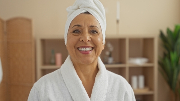 An older woman with a robe and a towel wrapped around her head smiles brightly.