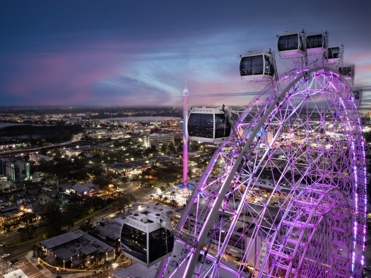 The top of a glowing ferris wheel with purple lights overlooking a glowing landscape of buildings at twilight.