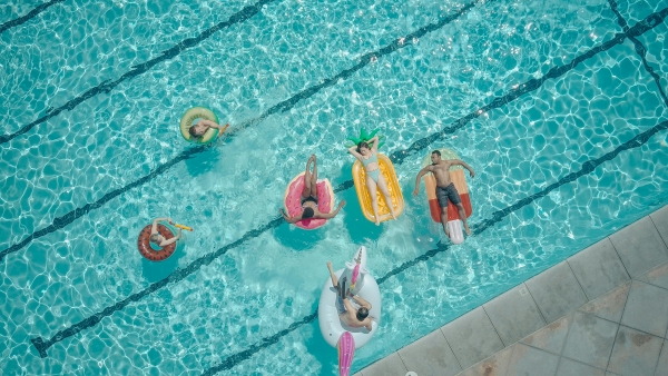 An overhead shot of six adults on whimsical pool floats lounging in a clear blue pool.
