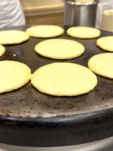 A close-up of rounded pancakes cooking on an iron griddle. 