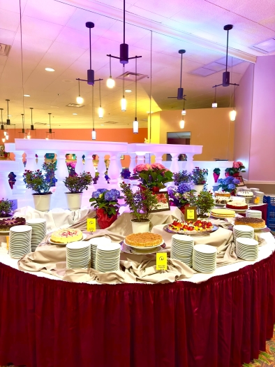 A large round table with a dark red skirt displaying various cakes and desserts, set with potted flowers and stacks of white plates. 