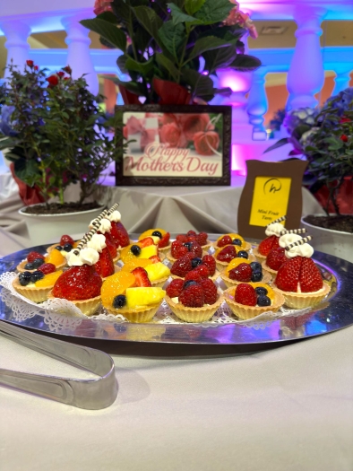 A silver tray displaying colorful mini fruit tarts with a variety of berries on a table with a sign that reads “Happy Mother’s Day” and several potted flowering plants. 