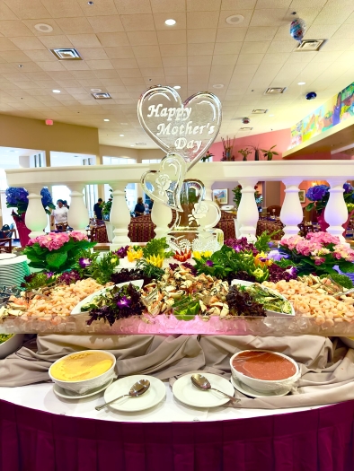 A display of fresh seafood featuring colorful flowers and greenery, as well as a large ice sculpture with hearts that reads “Happy Mother’s Day.” 
