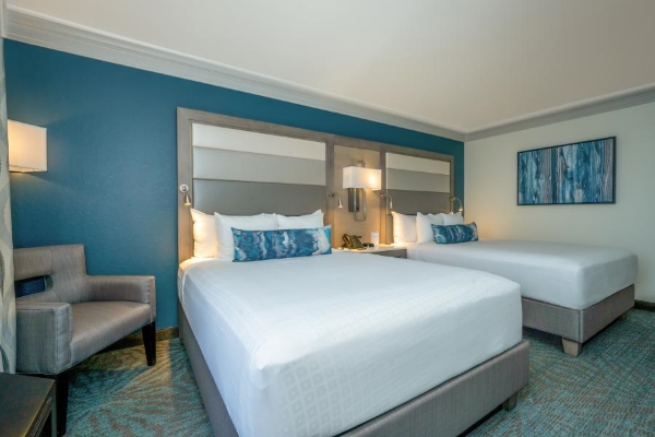 Two queen beds with white linens and blue throw pillows in a hotel room with a large chair, a blue accent wall, and warm lighting. 