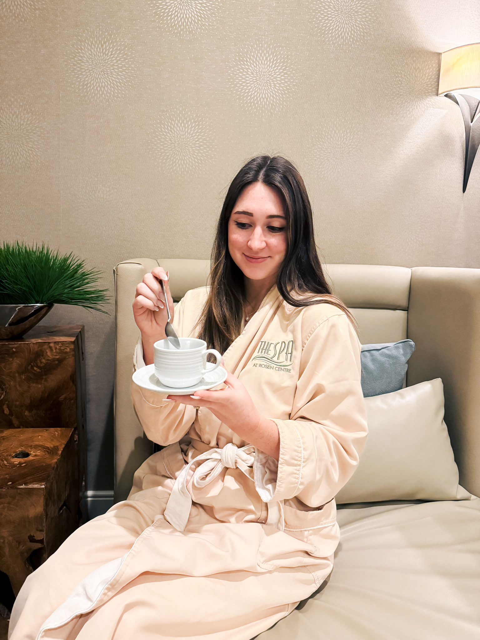 A brunette woman in a cream colored robe with The Spa at Rosen Centre logo on it stirs a mug of tea on a relaxing seat.