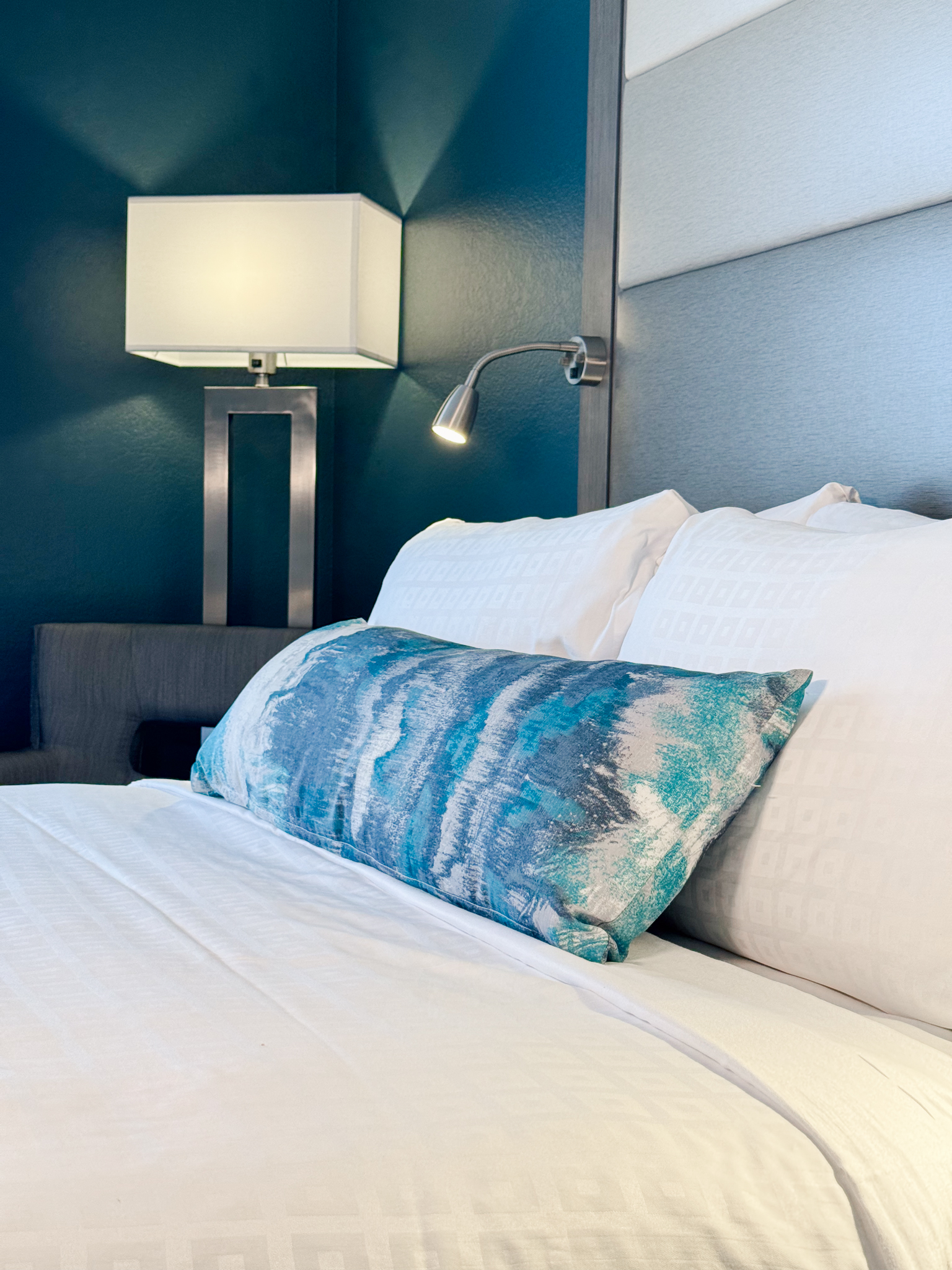 A close-up of a bed with white linens and a long blue throw pillow in a room with blue walls and a floor lamp.