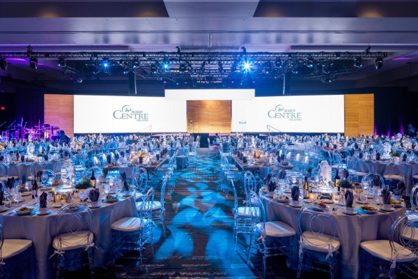 A large ballroom space with huge screens set up behind a stage at the front of the room and round tables set for a formal event. 