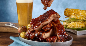 A table with a grey bowl full of BBQ ribs, a full glass of beer, and a plate of corn on the cob against a cloudy-blue gradient background. Rosen Centre Father's Day in Orlando.
