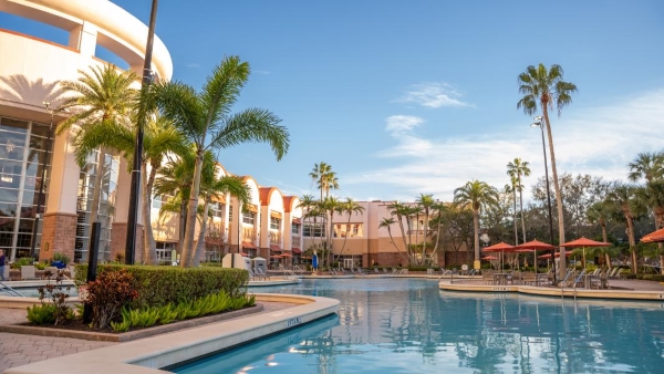 A view overlooking a clear blue pool with hotel arches, orange umbrellas, and palm trees. 
