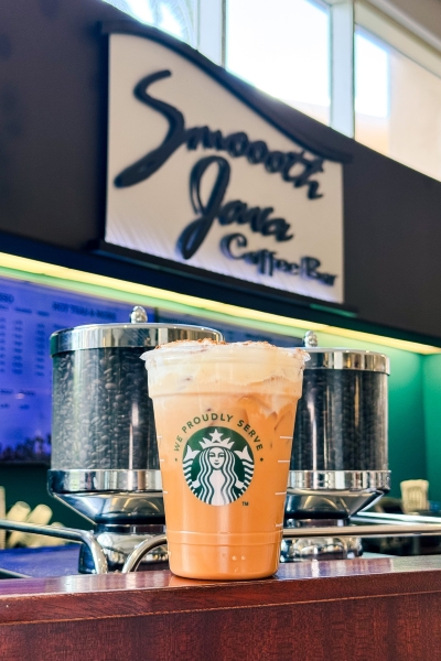 A full iced latte in a Starbucks cup sits on a counter in front of the Smoooth Java Coffee Bar sign at Rosen Plaza.