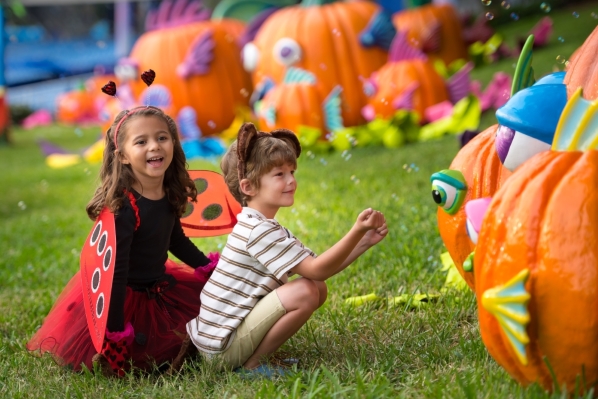 A boy and a girl in simple Halloween costumes kneel in grass in front of giant pumpkins decorated to look like fish.