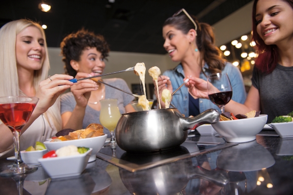 Four women around a table with a fondue pot, smiling and holding skewers of food dripping in cheese.