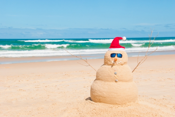 A snowman made of sand wearing a Santa hat and sunglasses on a sunny beach.