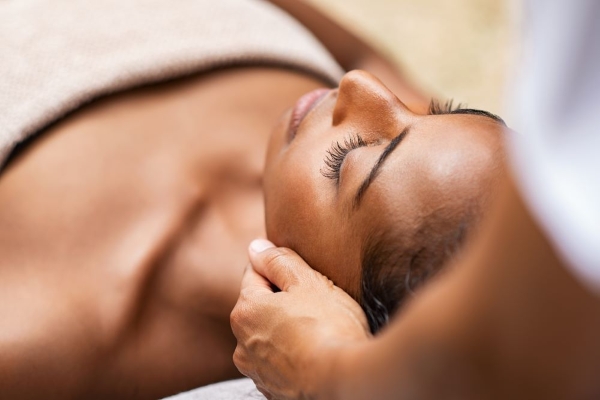 A woman rests with her eyes closed as a masseuse rubs her cheekbones.