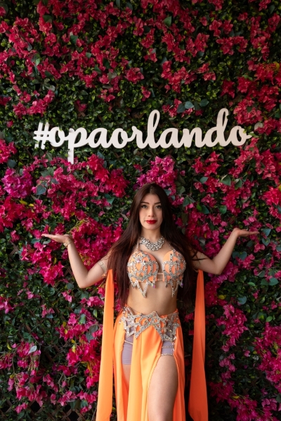 A woman in an orange belly dancing outfit stands in front of a floral wall with pink flowers and a sign reading #opaorlando.