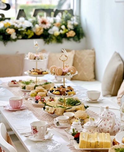 A table set with tiers of pastries and colorful teacups surrounded by pillows and flowers. 