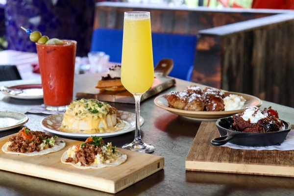 A table set with wooden boards of breakfast food, including tacos and eggs benedict, as well as a Bloody Mary and a mimosa.