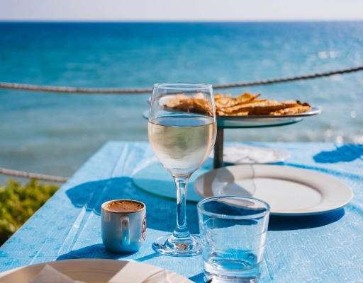 A close-up of a table set with a plate, a glass of wine, and a blue tablecloth overlooking an expanse of blue ocean water.