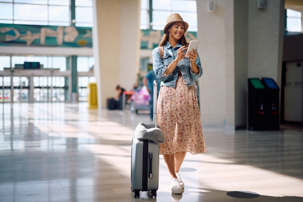 A woman in a dress and hat stands next to a rolling suitcase in a bright airport, looking at travel apps on her phone and smiling.