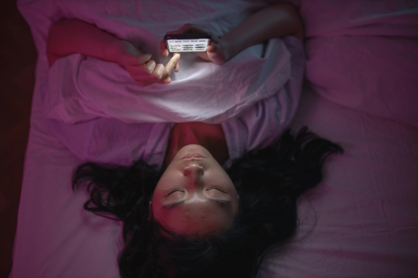 An overhead shot of a woman lying in bed in a dark room scrolling on a glowing phone.