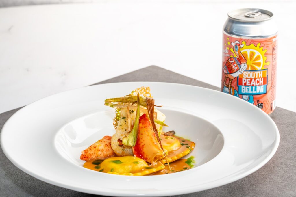 A bowl with two ravioli, lobster meat, and a garnish topper, and a can of beer in the background. 