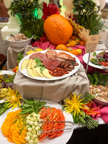 A selection of meats, cheeses, and tomatoes on a Thanksgiving buffet table decorated with pepper flower carvings, pumpkins, and herbs. 