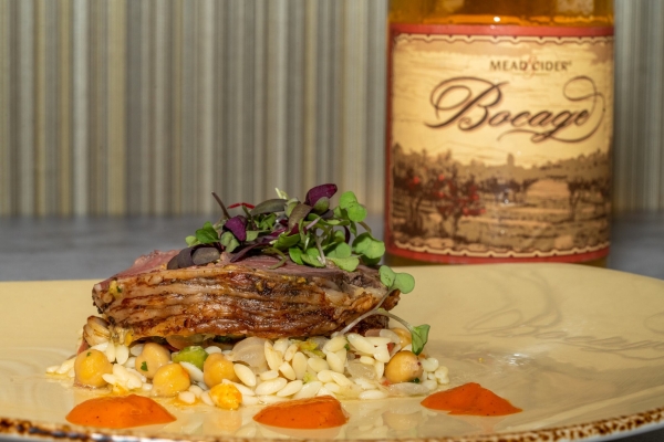 A cut of meat on a bed of orzo and chickpeas topped with microgreens, and a bottle with a vintage inspired label that reads Bocage mead cider standing behind it.