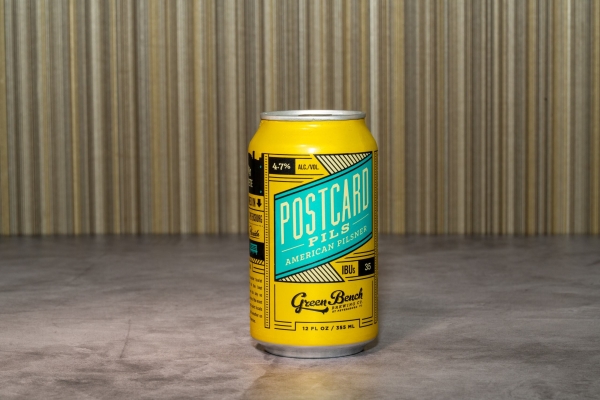 A yellow beer can with a teal banner across the center that reads Postcard Pils American Pilsner.