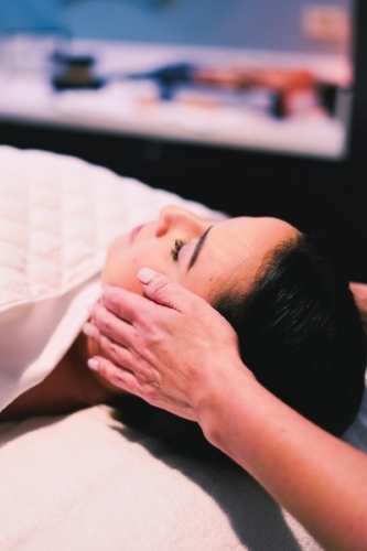A woman lies on a table covered in a blanket while hands massage her temples in a spa treatment.