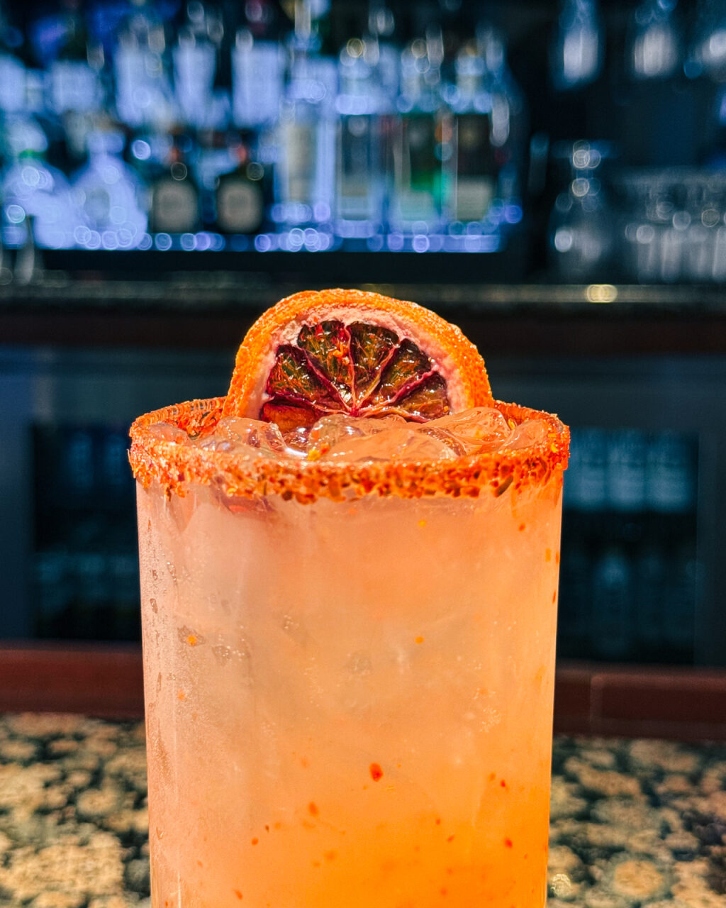 The top of a bright orange cocktail with a red tajin rim and a dried grapefruit garnish. 