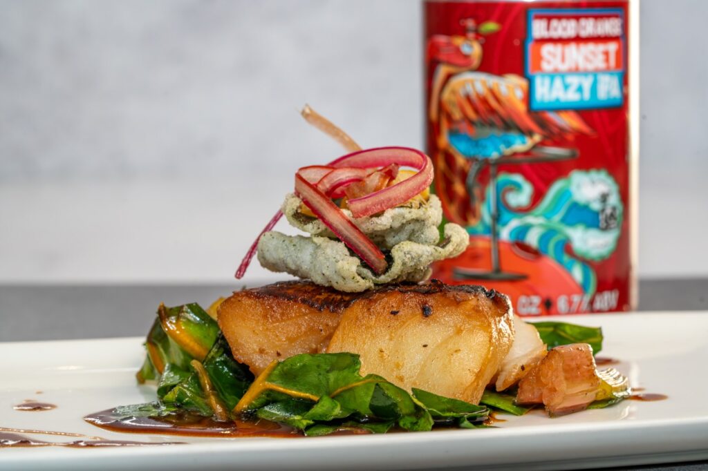 A fish filet on a bed of greens topped with a crisp and pickled onions and a can of beer in the background.