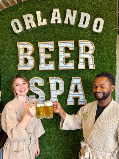 A man and woman smile and clink clear beer steins in front of a sign that reads Orlando Beer Spa.