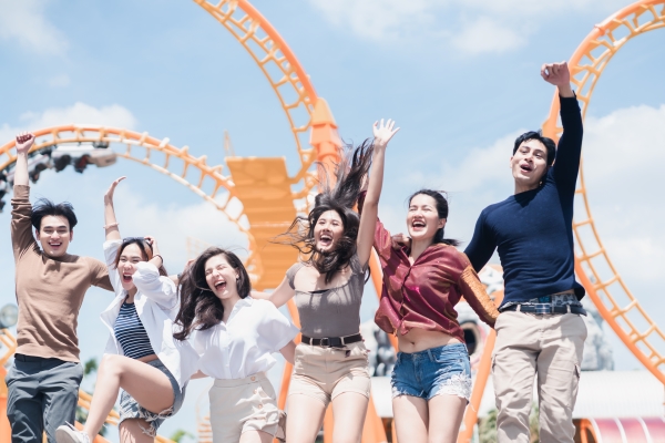 A group of six young adults jump in the air laughing in front of a yellow roller coaster track while on a friendcation.