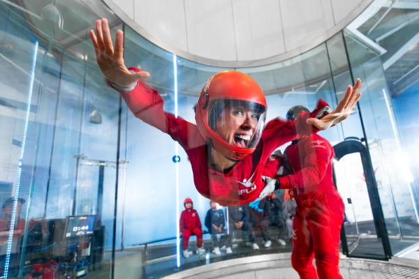 A woman in a red jumpsuit and helmet smiles while suspended during indoor skydiving, being guided by an instructor also in a red jumpsuit.