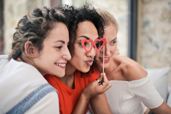 Three women in profile smile at a camera, while one holds a photo booth prop of heart-shaped glasses to her face.