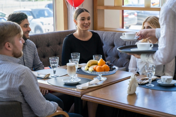  Two women and two men sit at a corner booth table set with a glass dish of fresh fruit, while a waiter sets coffee onto their table.