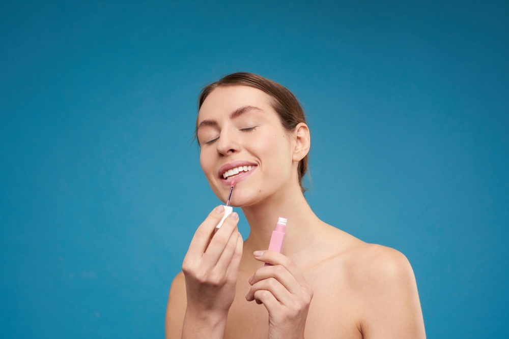 A woman stands against a blue backdrop, applying lip gloss to her mouth. 