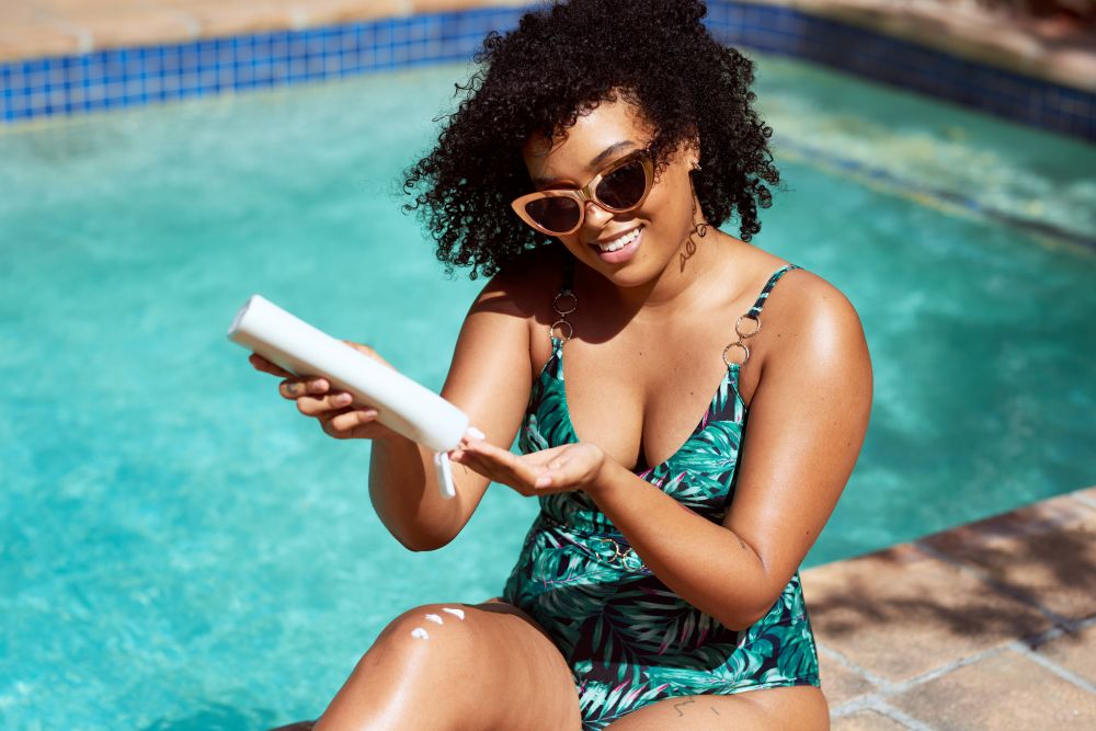 A woman in a green one-piece bathing suit and sunglasses sits by a pool applying sunscreen. Sunscreen is a must for summer skincare. 