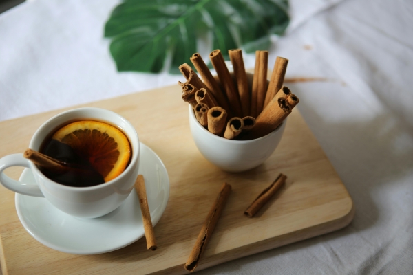 A white cup and saucer filled with tea and an orange slice beside a cup of cinnamon sticks on a wooden tray.