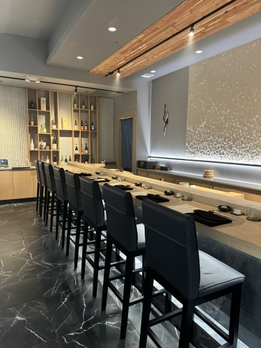 A restaurant with black marble floors and a long wooden bar with 8 tall stools seated before simple plate settings. 